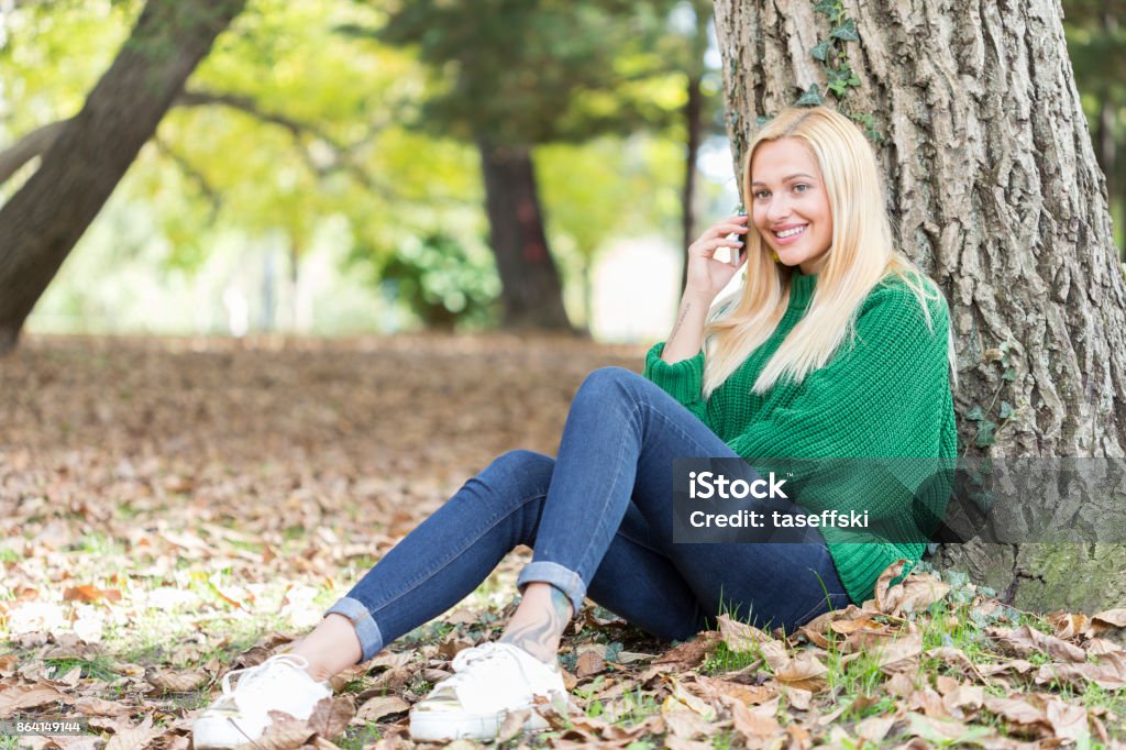 Smiling woman talking on her phone Happiness in Nature- Young woman sitting under tree and talking on phone Adult Stock Photo