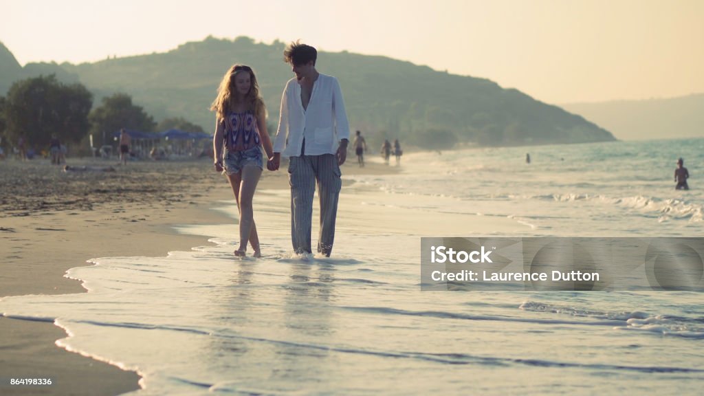 Beach walking towards A young heterosexual couple walking towards the camera along a beach lit by the late afternoon sun, the surf brushes there feet as they walk hand in hand along the sandy shore on vacation. Affectionate Stock Photo