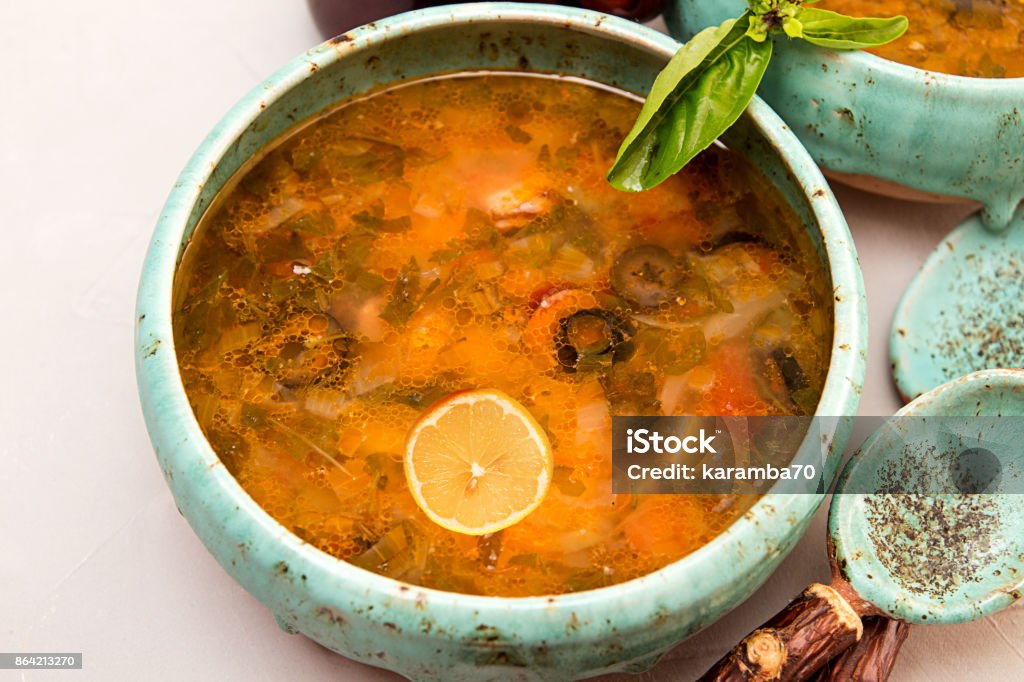 Soup with meat, smoked meat, sausages, pickled cucumbers, tomatoes, olives, herbs, lemon in blue bowl on gray concrete background, homemade food. Traditional Russian and Ukrainian soup solyanka. Backgrounds Stock Photo
