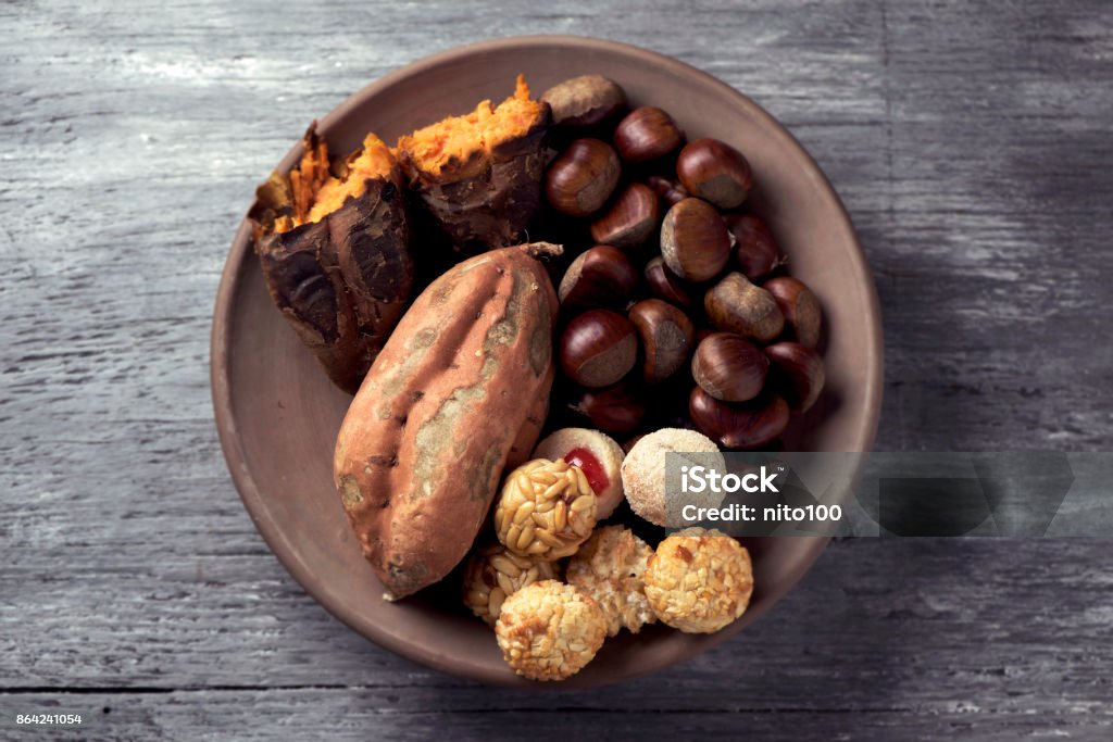 sweet potato, chestnuts and Catalan panellets high-angle shot of an earthenware bowl with some chestnuts, some panellets and a roasted sweet potato, as a typical snack eaten in All Saints Day in Catalonia, Spain, called castanada, on a table Castañada Stock Photo