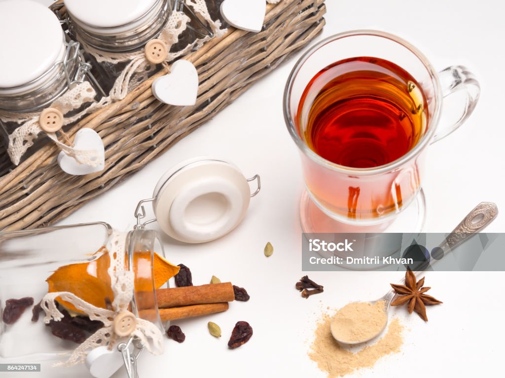 Tea with spices in a glass mug on a white background. Next basket with jars and spices: cinnamon, cardamom, orange, cloves. Spoon with ginger. Medical tea with spices in a glass mug on a white background. Next basket with jars and spices: cinnamon, cardamom, orange, cloves. Spoon with ginger. Basket Stock Photo