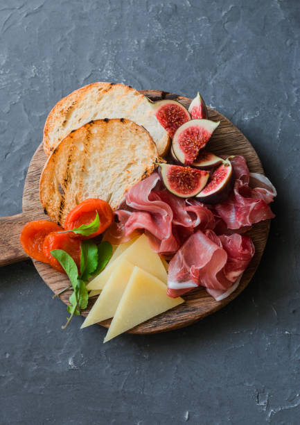 istock Delicious appetizers for wine or a snack - prosciutto, figs, bread, cheese on a rustic wooden cutting board. On a gray background, top view 864315656