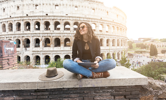 Woman sitting legs crossed in front of Coliseum and resting, using digital tablet. Wears casual clothes.