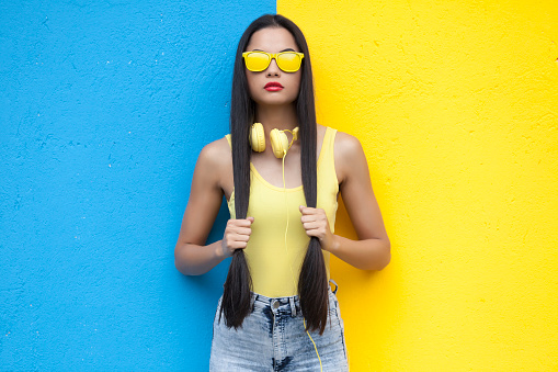 Woman wearing yellow vest, yellow headphones and holding her hair in front of yellow and blue wall background.