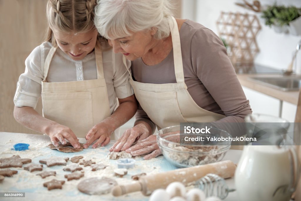 Happy granddaughter sculpting pastry from meal Creative girl is making form of cookies from dough. Her face is all in flour. Old woman is looking at her creation with satisfaction and smiling Grandparent Stock Photo