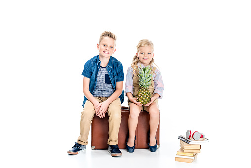 cute smiling kids with pineapple sitting on suitcase and looking at camera isolated on white