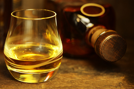 Close-up of a glass of whiskey on a rustic wooden table. A bottle of scotch whiskey can be seen in the background.