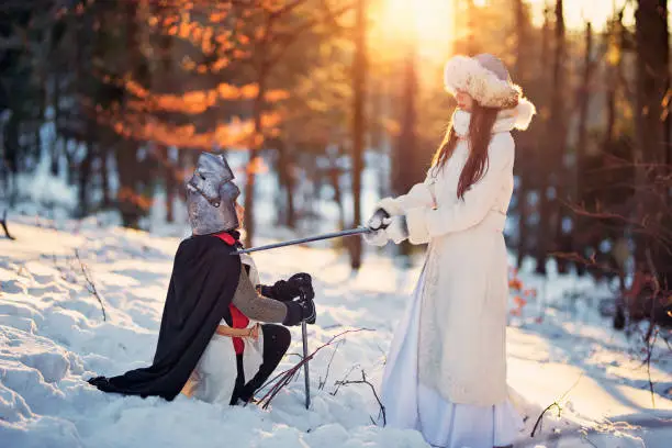 Young lady is knighting loyal knight with a sword. Snowy forest of a sunny winter day.
