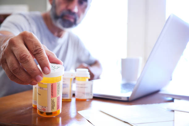 Hispanic Man Sitting At Dining Room Table Reaches For His Prescription Medications A Hispanic man in his late fifties reaches for one of his prescription medication bottles as he sits at his dining room table. His laptop computer is open in front of him while sunlight filters in through the window behind him bathing the room with a soft glow of light. Researching what Medication to buy stock pictures, royalty-free photos & images