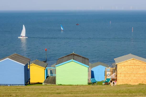Beach huts in Whitstable, kent, UK Colourful beach hits at the bottom of the grass area  in Tankerton, known as the slopes. The windfarm can be seen on the horizon herne bay stock pictures, royalty-free photos & images