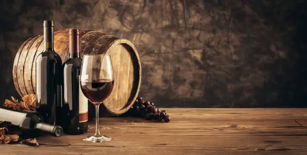 Wine glass, wooden barrel and collection of excellent red wine bottles in the cellar: traditional winemaking and wine tasting concept