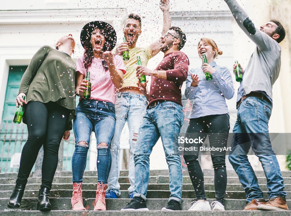 Happy crazy friends celebrating on the street drinking beer and throwing confetti - Young students having fun together - Friendship and youth concept - Vsco warm filter - Main focus on center guys Celebration Stock Photo