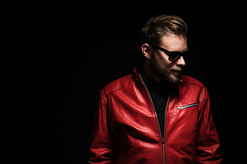 Cool man in a red leather jacket with sunglasses standing against a black background.