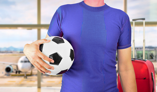 Cropped rear view image of a sports fan holding a soccer ball under his arm with blue t-shirt at airport