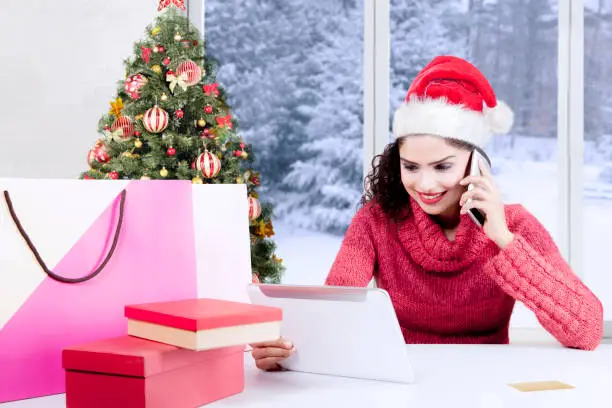 Beautiful woman purchasing Christmas presents online with a digital tablet computer while wearing a Santa hat and make a phone call at home