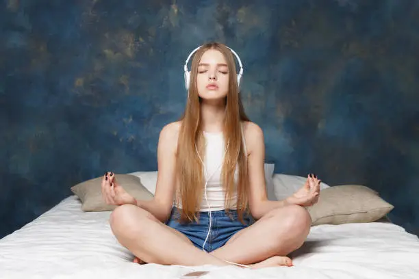 Beautiful girl meditates to the sound of music sitting on a bed with earphones