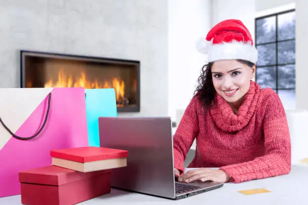 Pretty girl wearing Santa Claus hat and sitting at home while using a computer laptop for shopping online