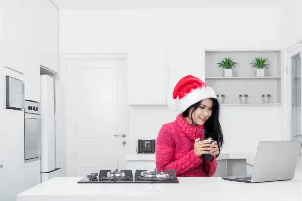 Pretty girl wearing Santa Claus hat while holding a mug of hot tea and looking at a laptop in the kitchen