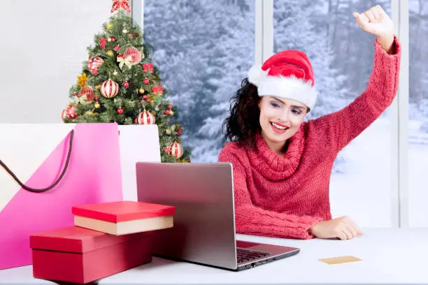 Pretty woman wearing Santa hat and sweater, shopping online Christmas gifts with laptop at home