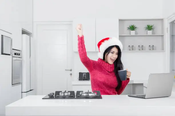 Portrait of successful pretty girl wearing Santa Claus hat while using a laptop and lifting her hand in the kitchen