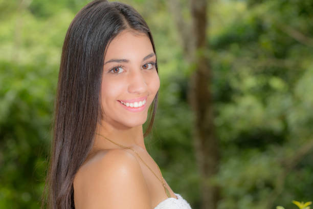 Beautiful latin young lady, teenager, poses for a headshot portrait on her side turnng her head to the camera. She is smiling.  Her beaufiful black long hair falls to her back. She is wearing a white strapless top. The background is of natural trees from park.