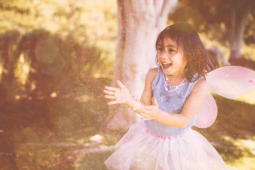 Little Asian girl in fairy costume playing and blowing glitter outdoors in park in spring