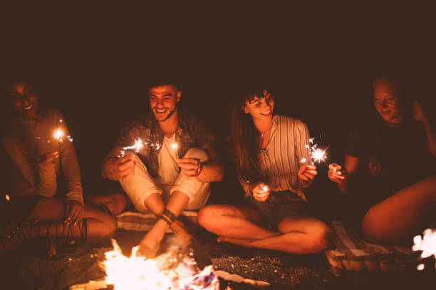 istock Multi-ethnic hipster friends celebrating with sparklers sitting around beach campfire 864534692