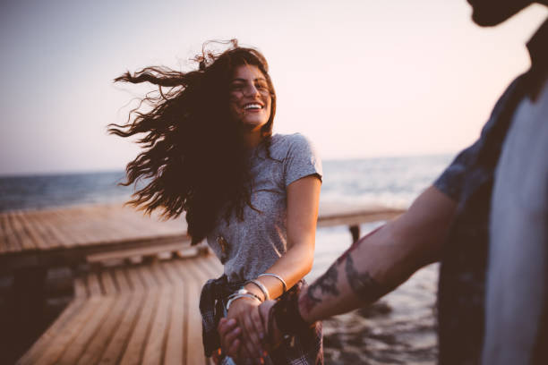 Photo of Young playful hipster couple holding hands on jetty at sunset