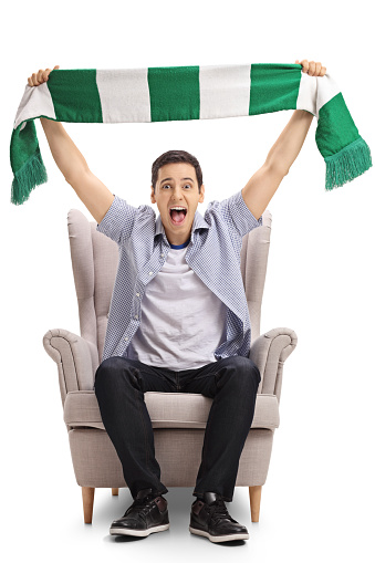 Excited sports fan sitting in an armchair and holding a scarf isolated on white background