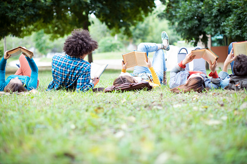 Hispanic teenager college student reading a book outdoors, laying on the grass