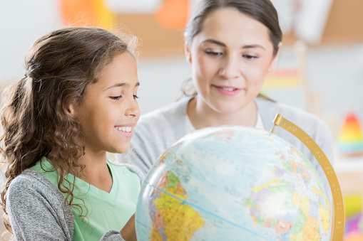 Curious mixed race elementary schoolgirl uses a globe to learn about geography at school. The girl's teacher is helping her find a country or continent on the globe.