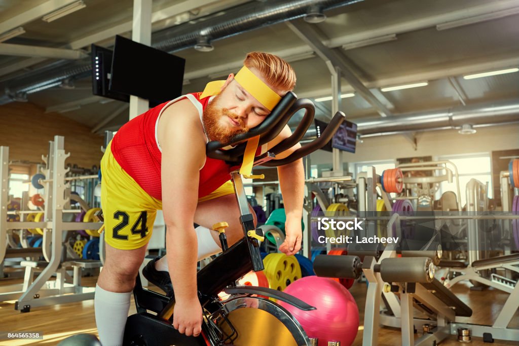 A fat man is tired on a simulator in the gym A fat man is tired on a simulator in the gym. Humor Stock Photo