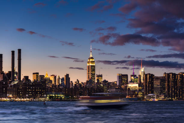 Sunset view of the iconic skyline of Lower Manhattan, from Williamsburg, Brooklyn. The East River Ferry is a blur of motion, and the Empire State Building dominates the cityscape.