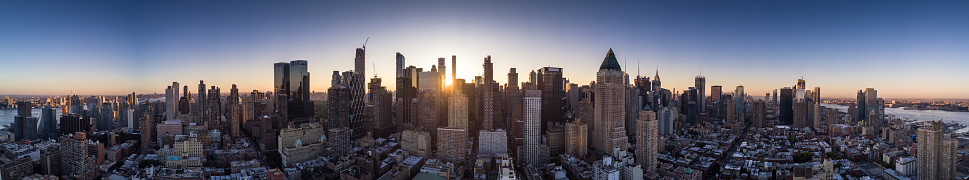 Panorama taken from a drone flying above Midtown Manhattan at dawn, with the sun about to rise behind the skyscrapers of the financial district.