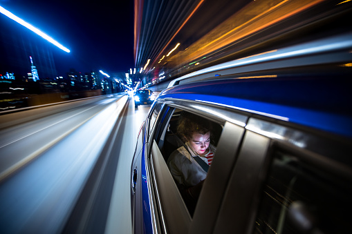 A woman sits in the back of a car, possibly a rideshare, looking at her mobile phone. The lights of New York City streak past as the car crosses the Brooklyn Bridge.