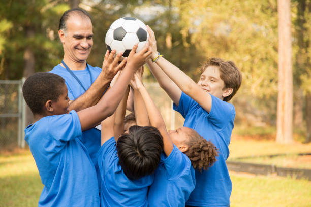 Children's soccer team coach celebrates victory with his players. Multi-ethnic and mixed age group of school children celebrate recent victory with their team soccer coach.  They all wear blue uniforms. youth sports competition stock pictures, royalty-free photos & images