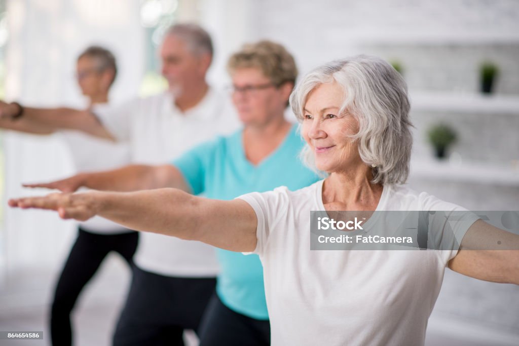 Reaching Out A multi-ethnic group of adult men and women are indoors in a fitness studio. They are wearing casual clothing while at a yoga class. A senior Caucasian woman is smiling while stretching out her arms. Senior Adult Stock Photo