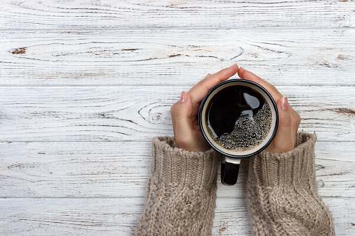 Woman holding cup of hot coffee on rustic wooden table, closeup photo of hands in warm sweater with mug, winter morning concept, top view.