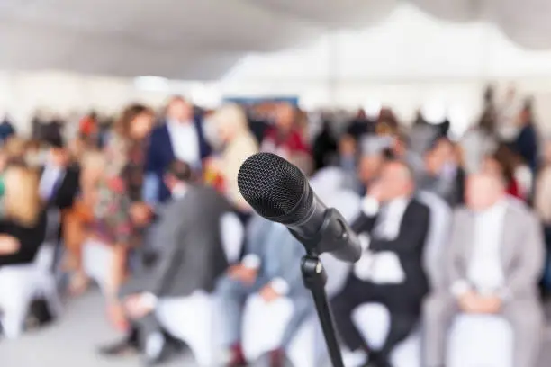 Photo of Microphone in focus against blurred audience. Participants at the business or professional conference.