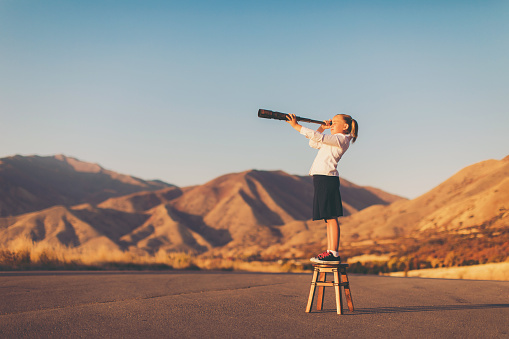 A young business girl or student dressed in business attire stands on a stool looking through a telescope. She is searching for her next big opportunity to prove her success and abilities. She is confident looking in the mountains of Utah, USA.