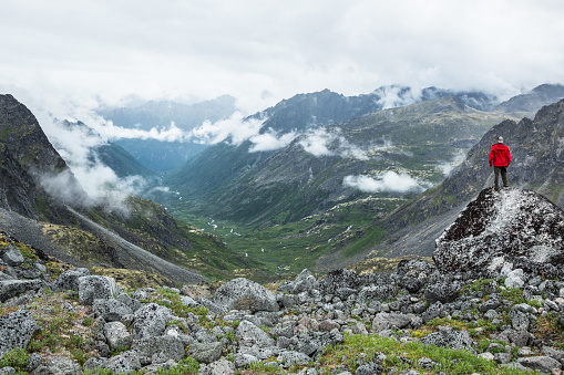 Young man in backward ballcap and red rain jacket stands overlooking a huge glacial valley with clouds lining it's edges and green plants filling the inside of the valley. He stands near the Snowbird Glacier looking out into the deep wilderness of the Talkeetna Mountain Range of Alaska