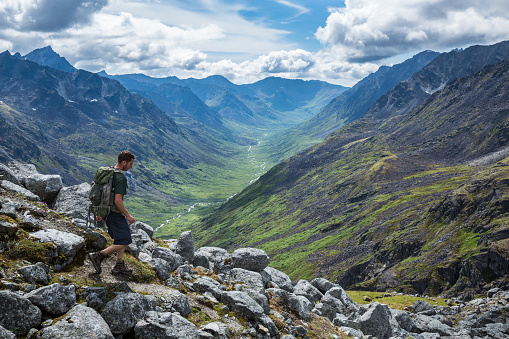 A man in his mid-20's hikes with a daypack down a steep trail overlooking the Little Susitna River in the Gold-Mint Trail Valley of the Hatcher Pass area in the Talkeetna Mountain Range of Alaska.