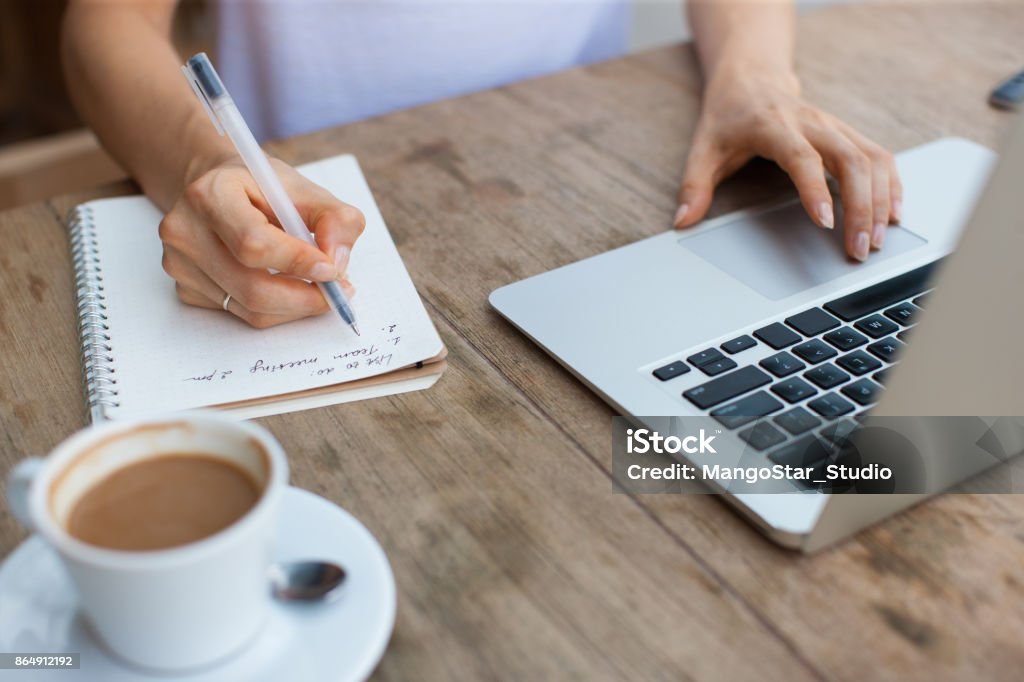 Cropped View of Woman Working on Laptop in Cafe Cropped view of business woman hands working, typing on laptop computer and making notes in notebook at cafe table List Stock Photo