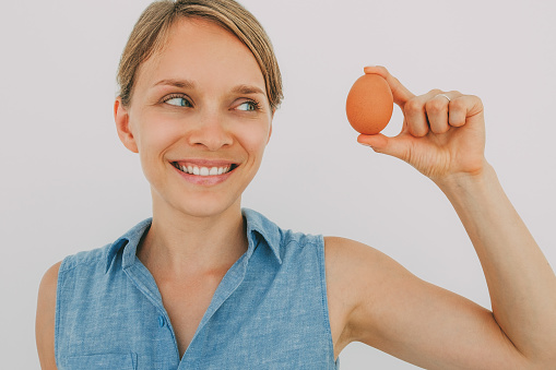 Closeup portrait of smiling young attractive woman holding egg with two fingers and looking at it. Isolated front view on grey background.