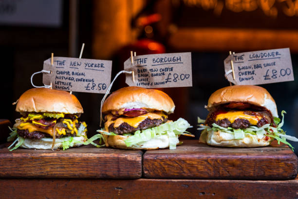 Freshly flame grilled burgers displayed in a row at Borough Market, London Close up image of a selection of freshly flame grilled burgers in a row on a wooden counter at Borough Market, one of the oldest and most famous food markets in the world. Each of the burgers has its own label, on which is written the contents of the burger. The burgers are sandwiched between glazed buns, and presented on beds of fresh green lettuce and stuffed with melted cheese and red onion. Horizontal colour image with copy space. borough market stock pictures, royalty-free photos & images