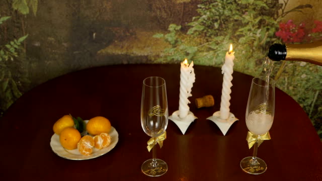 Pouring sparkling champagne wine into the glass against wooden background. Mandarins in plate on table. Light from pair candles. Evening indoors shot.