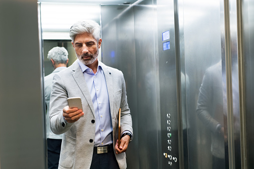 Mature businessman standing in the elevator holding smartphone, texting.