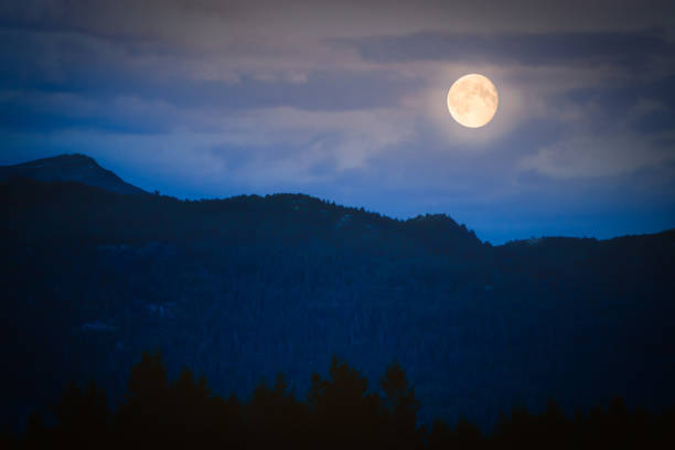 Full moon in dusk over dark mountains with clouds in background and forest in foreground