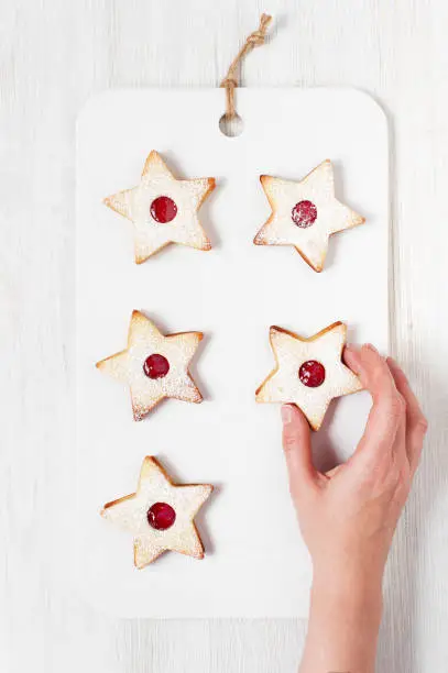 Female hand holding the traditional Linzer cookies - star with red jam on the white wooden table. Christmas and New Year pastry.