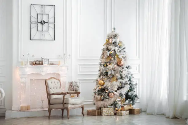 Classic white christmas interior with new year tree decorated. Fireplace with grey chair, clocks on the wall and presents under the tree